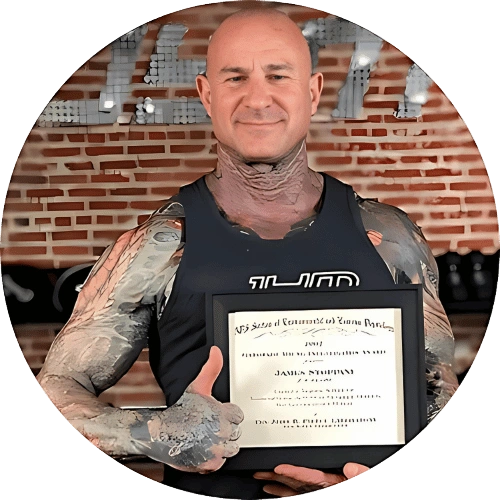 A muscular, tattooed man with a bald head is smiling and giving a thumbs up. He is holding a framed certificate and standing in front of a brick wall. The certificate appears to be an award from the "State of Council of Zone Body." He is wearing a sleeveless black shirt.