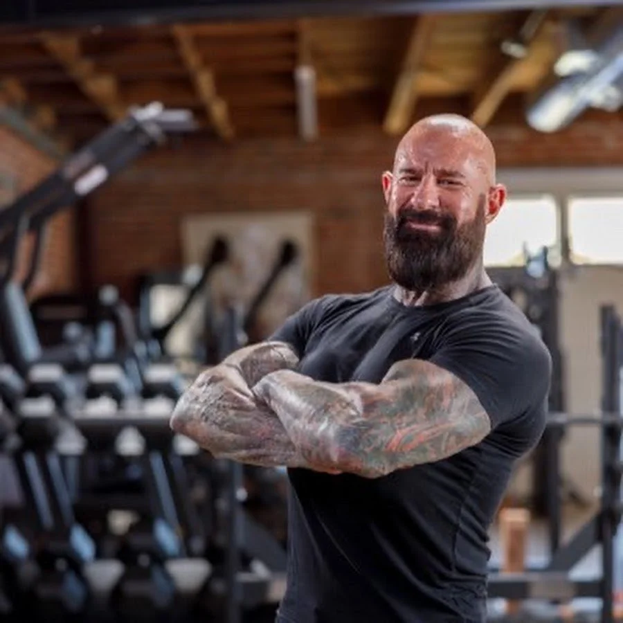 A man with tattoos on his arms, who happens to be a personal trainer inspired by Dr. Jim Stoppani, helps clients achieve their fitness goals.